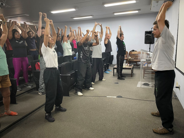 Pessoas vistas em um auditório com os braços para cima realizando a prática seguindo um professor